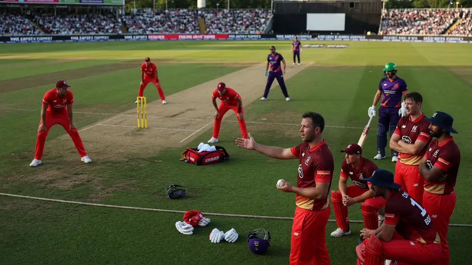 Medium shot of fielders and a batter setting for a tactical moment on a floodlit cricket ground